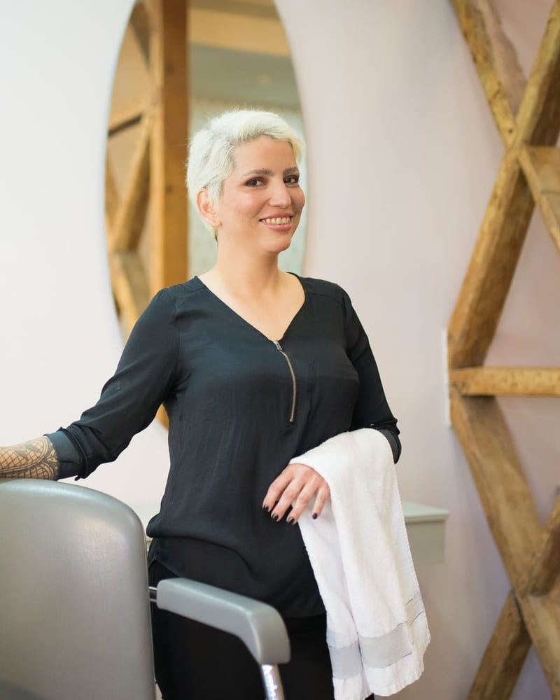 Sabine Bäuml lächelt im hellen Atelier, die Hand auf einem Friseurstuhl, ein Handtuch über dem Arm